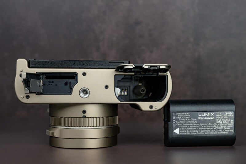 A digital camera shown from the bottom, with compartments for the SD card and battery open and the battery removed, revealing their ports. The camera body and battery are placed on a dark, blurred surface.