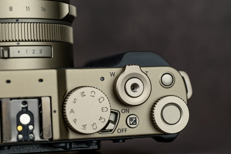 Close-up of a metallic camera’s top controls, showing dials for settings, mode selection, power switch, a button labeled "C," and part of the lens with visible focus and zoom markings.