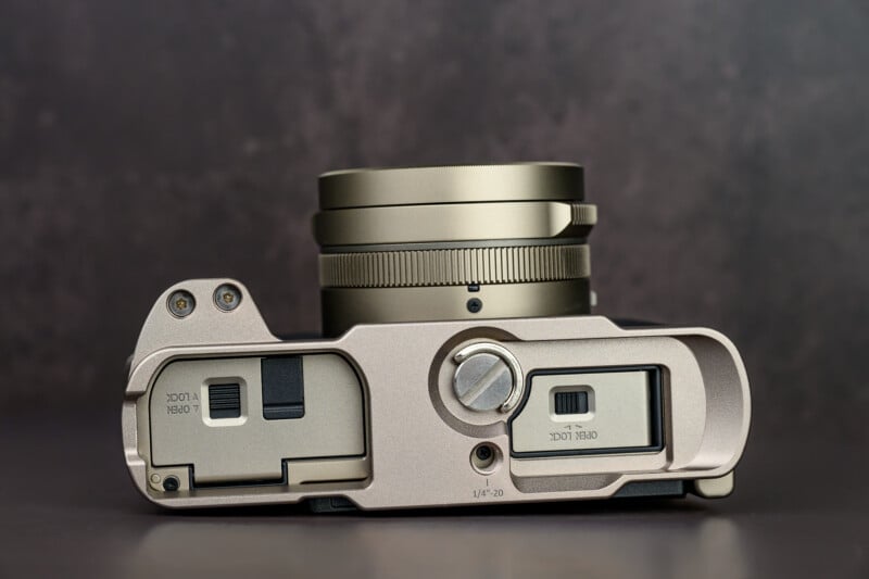 A close-up view of the top and bottom of a compact digital camera with a metallic finish, showing the lens, dials, and battery/memory card compartments against a dark background.