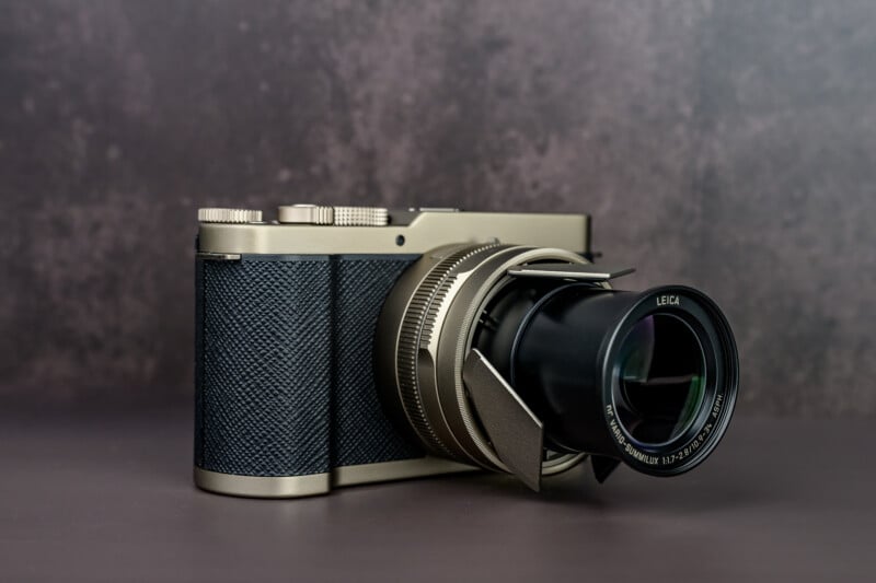 A silver and black Leica camera with a textured grip and extended zoom lens sits on a flat surface against a blurred, dark background.