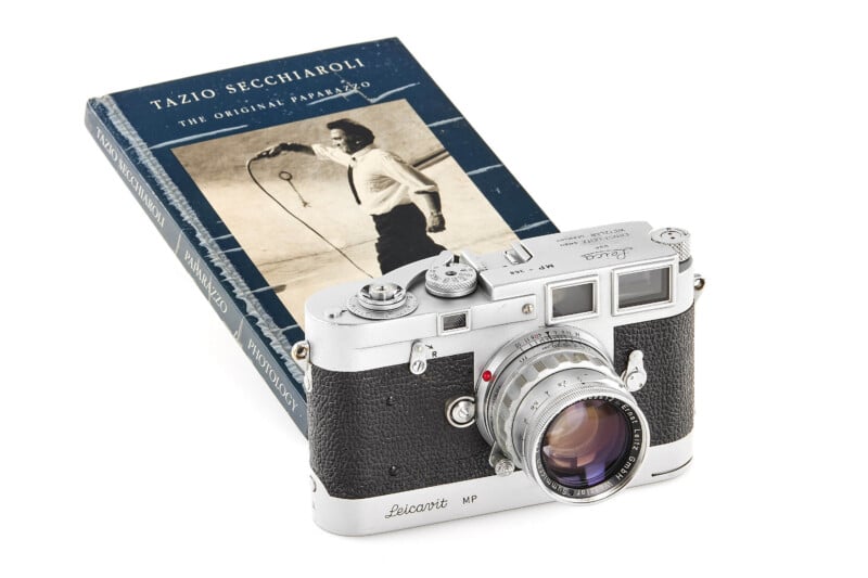 A vintage Leica camera rests on top of a book titled "Tazio Secchiaroli: The Original Paparazzo," featuring a black-and-white photo of a man with a camera on the cover.