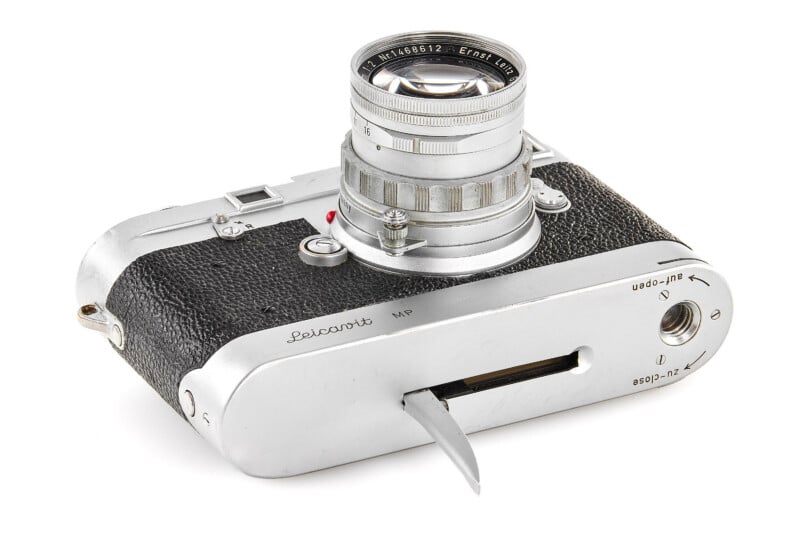 A vintage Leica camera with a black textured body, silver lens, and metal detailing, shown partially open with its film advance lever extended and a view of the bottom plate controls.
