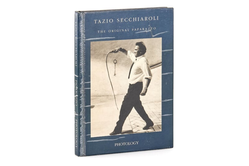 A hardcover book titled "Tazio Secchiaroli: The Original Paparazzo" features a black-and-white cover photo of a man holding a coiled cord, with a blue and white border. The publisher, Photology, is noted at the bottom.