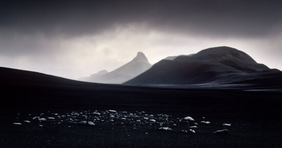 A dramatic, dark landscape with rocky hills and a prominent jagged peak in the background. Foreground features scattered stones on a stark, shadowy plain under a moody, cloudy sky with light breaking through.