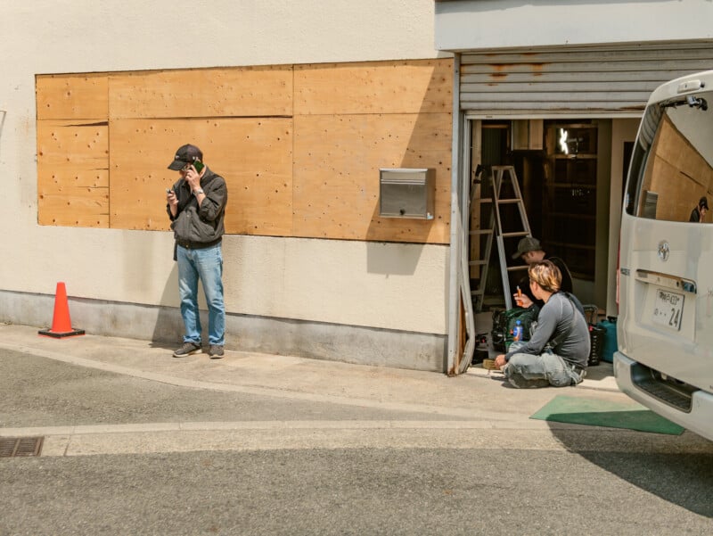 A man stands by a wall talking on the phone near a boarded-up window, while two people sit on the ground inside a partially open garage, surrounded by tools and a ladder. A white van is parked nearby.