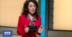 A woman with curly brown hair, wearing a red blazer and patterned scarf, stands indoors holding a black electronic device. The background is yellow with a blue vertical column. BBC Archive logo appears in the bottom left corner.