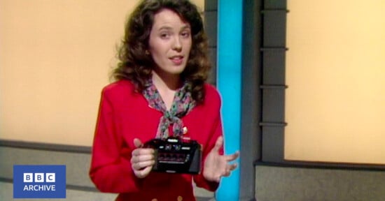 A woman with curly brown hair, wearing a red blazer and patterned scarf, stands indoors holding a black electronic device. The background is yellow with a blue vertical column. BBC Archive logo appears in the bottom left corner.