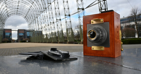 A vintage wooden camera sits on a metal surface outdoors, with film holders next to it. In the background, there is a large metal arch structure and modern buildings, under a cloudy sky.