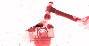 A hammer is striking an old Praktica film camera, causing it to break apart with debris and small pieces flying, all shown with a red tint.