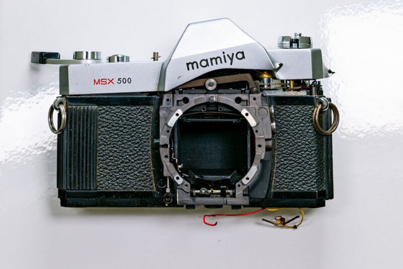 A disassembled vintage Mamiya MSX 500 film camera with the lens removed, revealing the inner mount and mechanical parts, lying on a white surface.