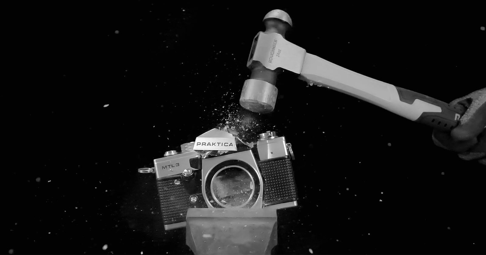 A hammer is smashing an old Praktica MTL3 camera, shattering its lens and body, with debris flying against a black background.