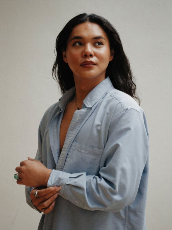 A person with long dark hair wearing a light blue button-up shirt looks to the side with a slight smile, standing against a plain light-colored background.