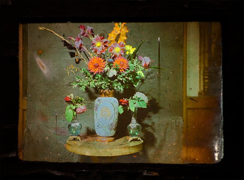 A colorful bouquet of assorted flowers in a decorated blue vase sits on a round table, flanked by two smaller glass vases each holding a single flower, against a muted green background.