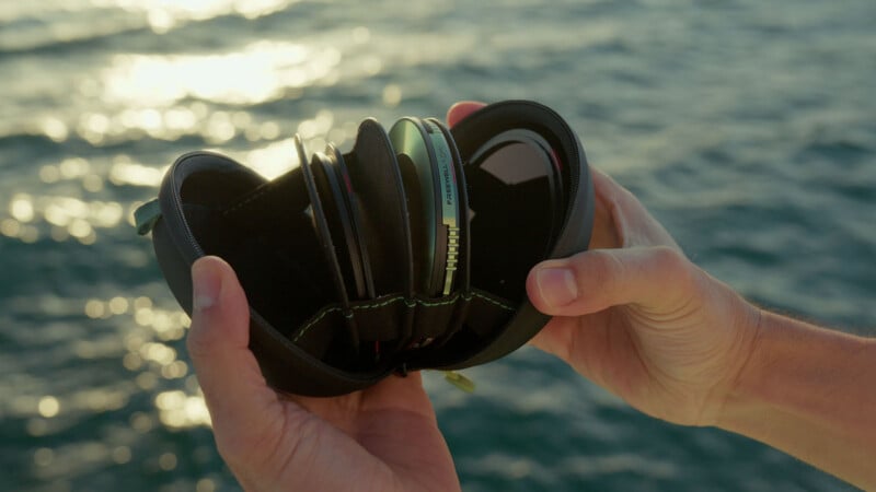 A person holds an open black zippered case containing several camera lens filters, with sunlight glinting off the ocean water in the background.