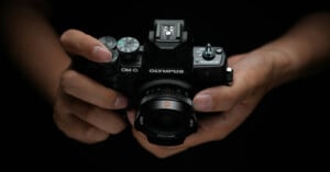 A person holds an Olympus OM-D digital camera with both hands against a dark background, focusing on the camera and the person's fingers on the controls.