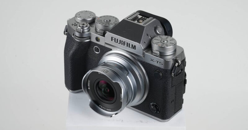 A Fujifilm X-T5 mirrorless digital camera with a black textured grip and silver top, equipped with a Fujinon lens, is displayed on a white platform against a plain background.