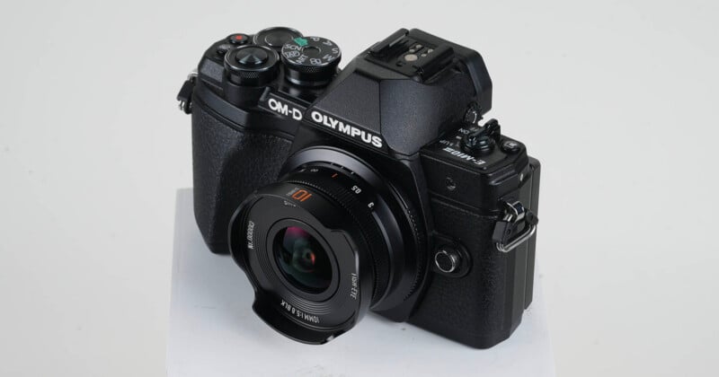 A black Olympus OM-D camera with attached lens is displayed on a white surface against a plain light background. The camera has various control dials and buttons visible.