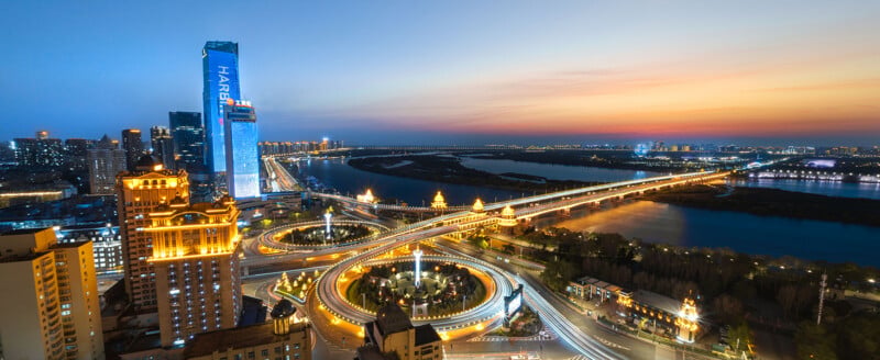 A cityscape at dusk featuring a brightly lit skyline, a tall building displaying "HAIER," intertwining highways, and bridges spanning a wide river under a colorful sunset sky.