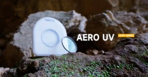 A camera lens filter with its case sits on a mossy rock in front of a rocky, cave-like background. Text reads "AERO UV" and "APEX SERIES.