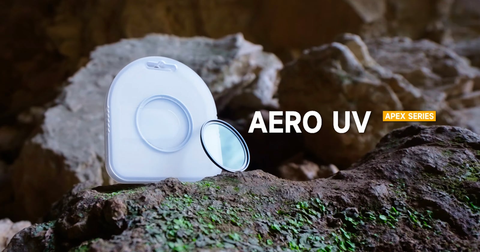 A camera lens filter with its case sits on a mossy rock in front of a rocky, cave-like background. Text reads "AERO UV" and "APEX SERIES.