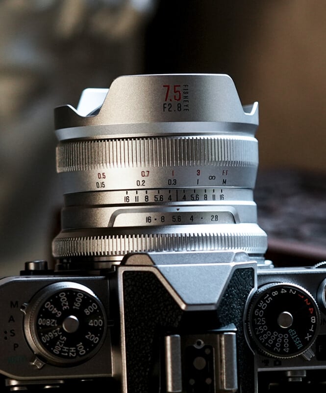 Close-up of a silver 7.5mm f/2.8 camera lens mounted on a vintage film camera body, showing detailed focus and aperture markings, with dials and buttons visible.