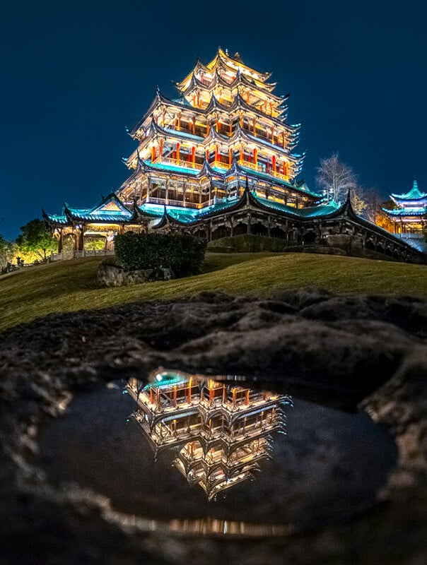 A brightly lit traditional Asian pagoda at night is reflected in a small puddle on the ground, with the structure’s colorful lights contrasting against the dark sky.