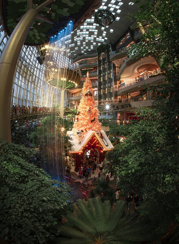 A large, white Christmas tree with lights and ornaments stands inside a lush indoor garden, surrounded by greenery, a small house, and cascading water features beneath a glass atrium ceiling. People are gathered around the display.