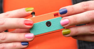 A person with colorful, mismatched nail polish holds a small, retro-style toy camera with an orange and teal body against an orange background.