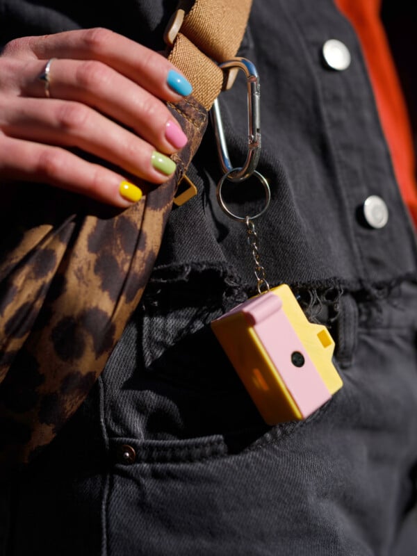 A person with multi-colored nails holds a leopard-print bag with a beige strap. A small, yellow and pink keychain camera is attached to the bag. The person wears a black denim outfit.