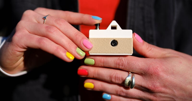 A person with brightly colored nails holds a small beige and white device with a button on top and a circular opening on the front, possibly a camera or electronic gadget.