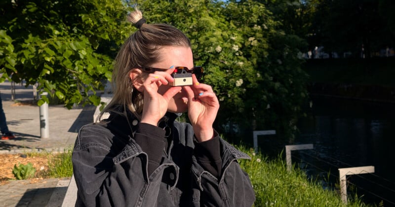 A person with light hair, wearing a dark jacket, stands outdoors by a canal and holds a small camera or viewfinder up to their eyes, surrounded by trees and greenery.