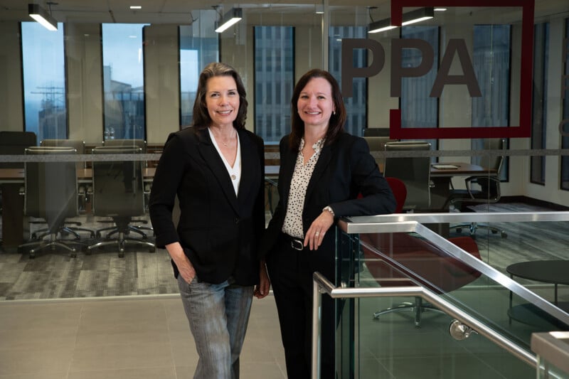 Two women in business attire stand smiling in a modern office with glass walls, desks, and large windows. A red-framed sign with the letters "PPA" is visible on the glass wall behind them.