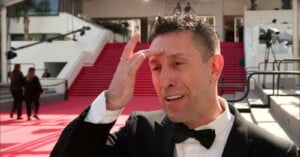 A man in a tuxedo gestures to his forehead while speaking outside on a red carpet, with large red stairs and a few people in the background.