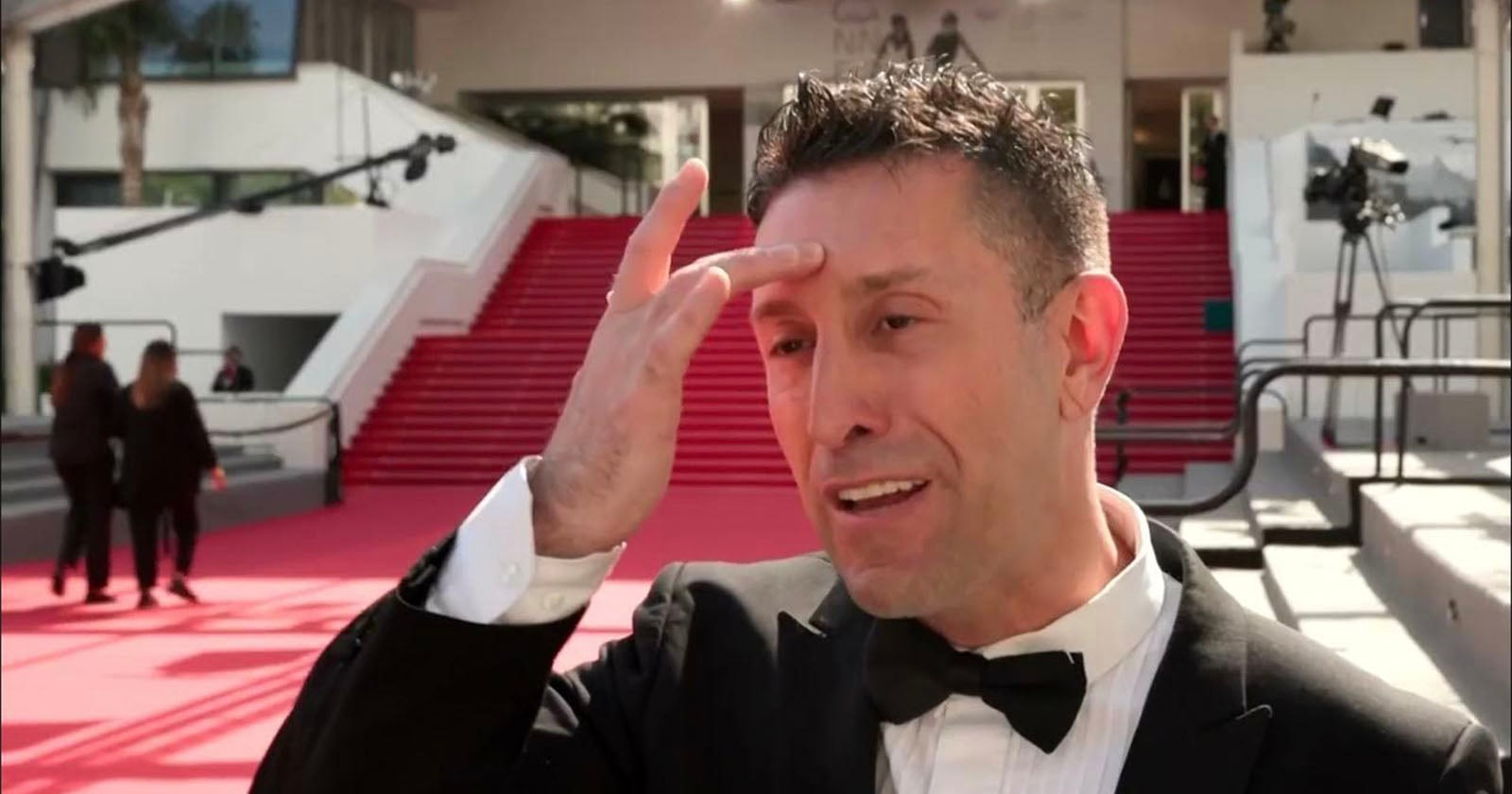 A man in a tuxedo gestures to his forehead while speaking outside on a red carpet, with large red stairs and a few people in the background.