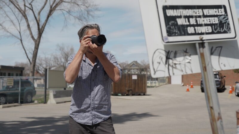 A person wearing a checkered shirt is taking a photo with a camera in a parking lot. A sign in the foreground reads, "Unauthorized vehicles will be ticketed or towed." Trees and buildings are visible in the background.