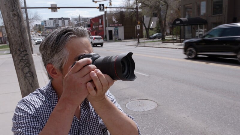 A person with gray hair and a checkered shirt stands on a city sidewalk, holding a camera up to their face and taking a photo. Cars are passing by on the street in the background.