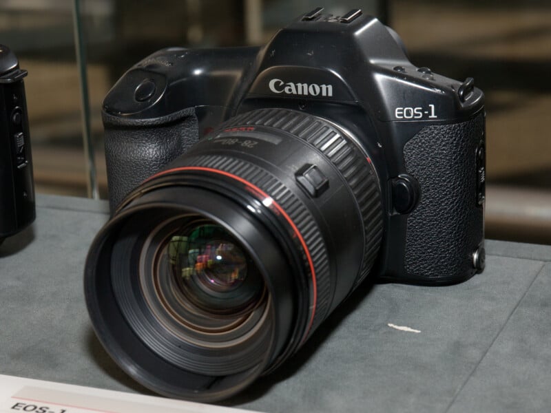 A Canon EOS-1 camera with a large lens attached is displayed on a flat surface, shown from a front angle. The camera body is black with visible brand markings.