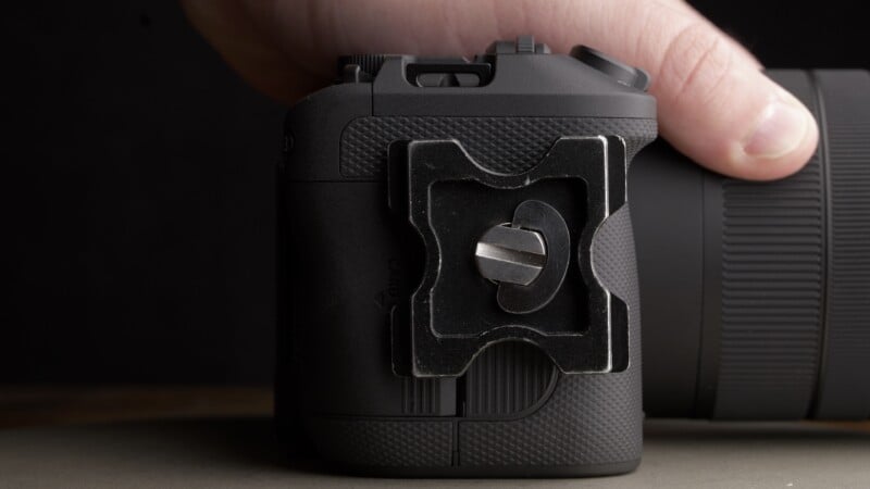 A close-up of a person’s hand holding a black camera with a metal tripod mounting plate attached to its side. The camera lens and mounting plate are clearly visible.