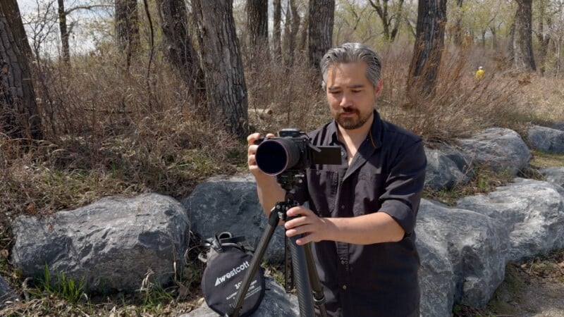 A man with gray hair and a beard adjusts a camera on a tripod outdoors, standing on rocks near trees and dry grass. A black camera bag rests on the rocks beside him.