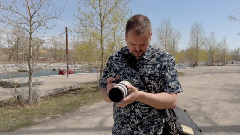 A man wearing a patterned shirt stands on a sunny park path, looking down at a camera with a large lens. He has a camera bag strap across his shoulder and trees without leaves are visible in the background.