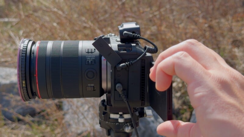 A close-up of a hand adjusting the LCD screen on a digital camera with a large lens mounted on a tripod outdoors, with dry brown grass in the background.