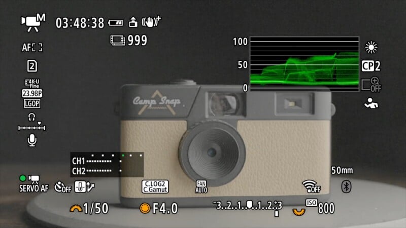 A digital camera screen displays a beige and gray film camera, with various camera settings and a green waveform monitor overlayed, showing exposure and focus information.