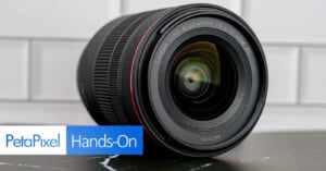 A close-up of a Canon RF 20-50mm F4 L IS USM camera lens resting on its side on a black surface, with a white tiled background. “PetaPixel Hands-On” text is overlaid in the bottom left corner.