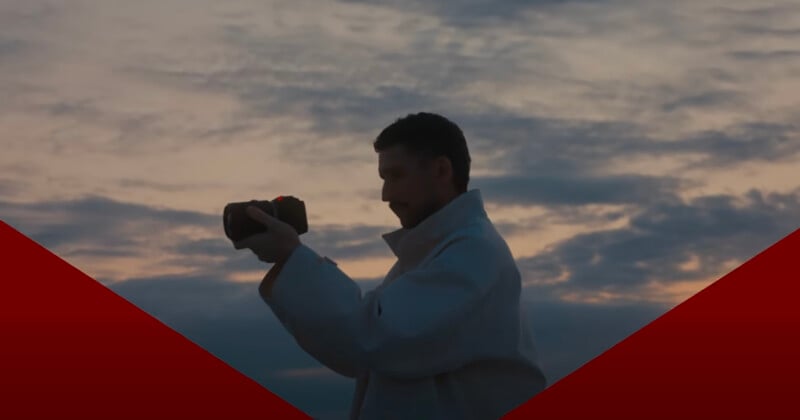 A man in a white jacket holds a camera, taking a photo or video at sunset with a cloudy sky in the background. Red geometric shapes frame the bottom corners of the image.