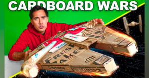 A man in a red hoodie holds a cardboard spaceship made from pizza boxes, styled like the Millennium Falcon. The background is split between green and a space scene with a small TIE fighter. Text reads "CARDBOARD WARS.