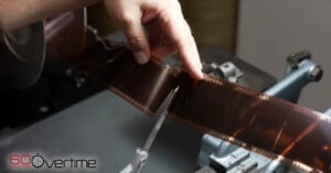 A hand uses scissors to cut a strip of photographic film on a worktable. The “60 Minutes Overtime” logo appears in the lower left corner.