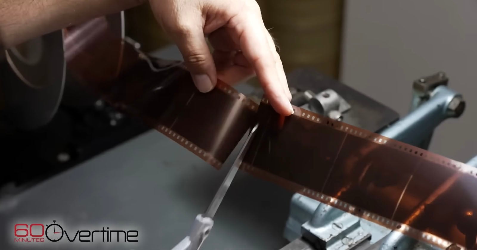 A hand uses scissors to cut a strip of photographic film on a worktable. The “60 Minutes Overtime” logo appears in the lower left corner.