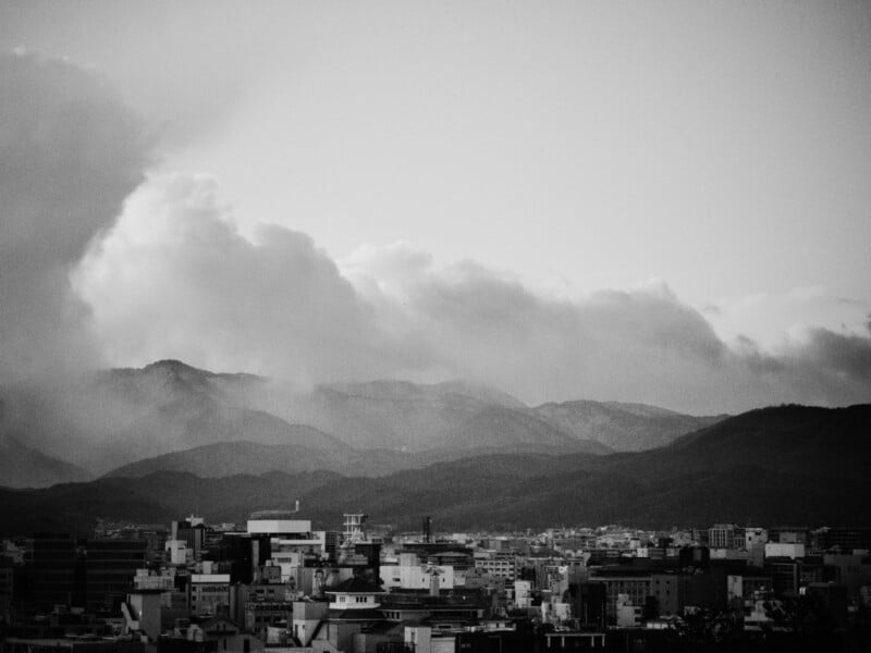 Black and white photo of a cityscape with buildings in the foreground and layers of mountains in the background, beneath a sky filled with dramatic, rolling clouds.