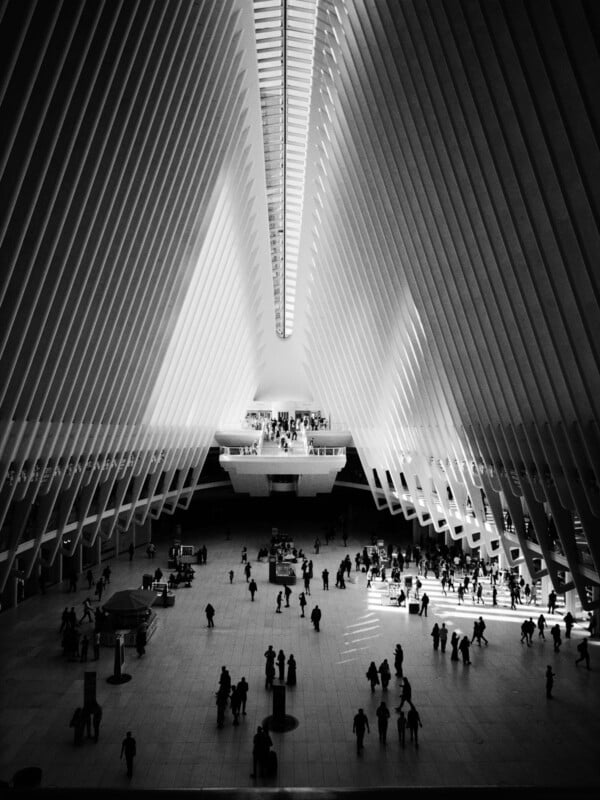 Black and white photo of a large, modern indoor space with tall, ribbed arches and a skylight. Silhouetted people walk across the open floor, casting long shadows in the dramatic light.