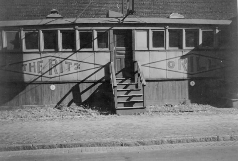 A vintage photo of a diner named "The Ritz Grill" shows a wooden building with multiple windows, a central staircase, and signs on either side of the entrance. The sidewalk and street are visible in the foreground.